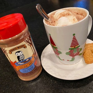 May include: A red jar of Barred Woods Maple Sugar Cocoa Mix with an owl logo. The jar is on a black surface next to a white mug with a red and green elf design filled with hot chocolate and topped with marshmallows. A gingerbread cookie is on a white saucer next to the mug.