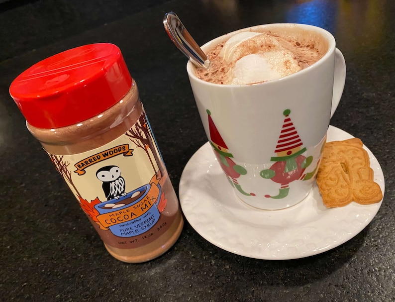 May include: A red jar of Barred Woods Maple Sugar Cocoa Mix with an owl logo. The jar is on a black surface next to a white mug with a red and green elf design filled with hot chocolate and topped with marshmallows. A gingerbread cookie is on a white saucer next to the mug.