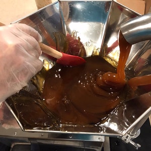 K&ouml;nnte beinhalten: Ein Trichter aus Edelstahl mit einem Auslauf gie&szlig;t eine dicke, braune Fl&uuml;ssigkeit in eine Edelstahlsch&uuml;ssel. Eine behandschuhte Hand r&uuml;hrt die Fl&uuml;ssigkeit mit einem Holzl&ouml;ffel und einem roten Spatel um.