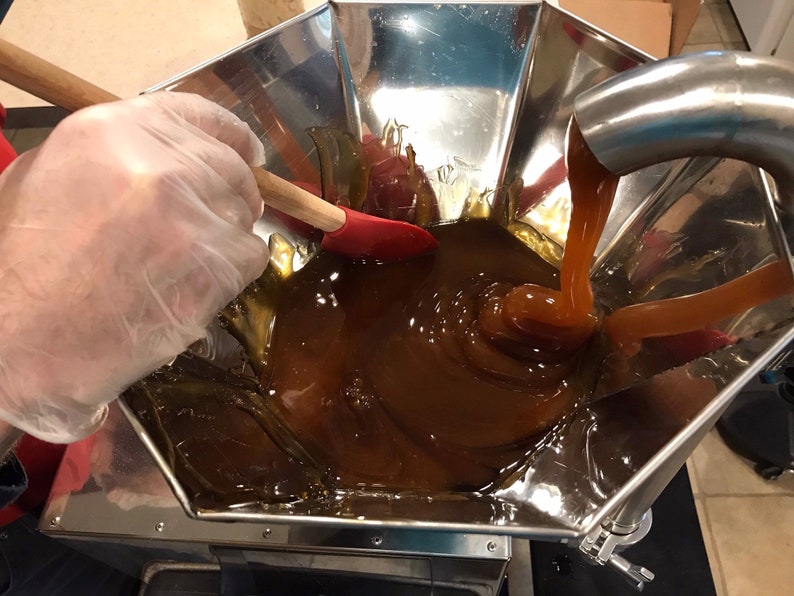 K&ouml;nnte beinhalten: Ein Trichter aus Edelstahl mit einem Auslauf gie&szlig;t eine dicke, braune Fl&uuml;ssigkeit in eine Edelstahlsch&uuml;ssel. Eine behandschuhte Hand r&uuml;hrt die Fl&uuml;ssigkeit mit einem Holzl&ouml;ffel und einem roten Spatel um.