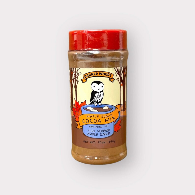 May include: A brown jar with a red lid containing maple sugar cocoa mix. The label features an illustration of an owl and a mug of hot cocoa. The text on the label reads "Barred Woods Maple Sugar Cocoa Mix Handcrafted with Pure Vermont Maple Syrup Net Wt. 12 oz. 340g"