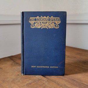Puede incluir: Un libro de tapa dura azul con letras doradas en el lomo y la portada. El título es "The Standard Operas" y el subtítulo es "New Illustrated Edition".