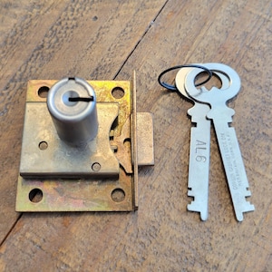 May include: A metal cabinet lock with two keys. The lock has a rectangular base with four holes and a cylindrical lock mechanism. The keys are silver-colored with the text "AL6" and "CORBIN CABINET LOCK DIV. NEW BRITAIN, CONN. U.S.A."