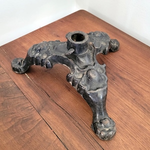 May include: A black, ornate metal stand with a central cylindrical opening and three decorative legs. The legs have a claw-foot design and are connected by a detailed, sculpted base. The stand is likely for a table or other furniture.