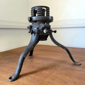May include: A dark, weathered cast iron Christmas tree stand with three curved legs and a central cylindrical holder. The metal surface shows signs of oxidation and wear. The text "PATENT APPLIED FOR" is clearly embossed on the central housing.