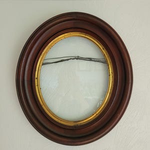 May include: An oval-shaped picture frame featuring a dark brown wooden outer border and a thin, inner gold-toned rim. The frame includes a clear glass pane and a dark wire stretched horizontally across the center for hanging purposes.