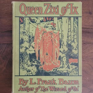 May include: A vintage book titled "Queen Zixi of Ix" by L. Frank Baum, author of "The Wizard of Oz." The cover features an illustration of a woman in a red dress with a forest backdrop. The book has a light green cover with orange lettering.