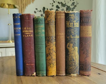 Könnte beinhalten: Eine Sammlung von sieben antiken Hardcover-Büchern, die aufrecht stehen und in verschiedenen Farben wie Blau, Kastanienbraun, Grün, Gelb und Braun gehalten sind. Die Buchrücken zeigen goldene und schwarze Prägeschriften sowie dekorative Muster mit Titeln wie Eminent Americans, Caesar Interlinear, Shadows and Sunbeams, Life of Lincoln, Human Body, The War Trail und Revised New Testament.