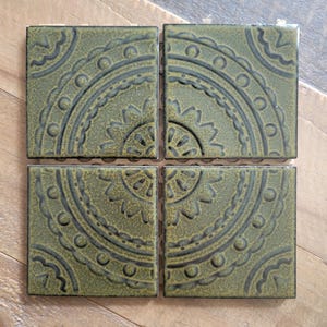 May include: Four square ceramic tiles arranged in a grid, each featuring a deep olive green glaze with a dark, recessed embossed pattern. The design on each tile forms a quarter of a circular mandala motif with intricate lines, dots, and petals.