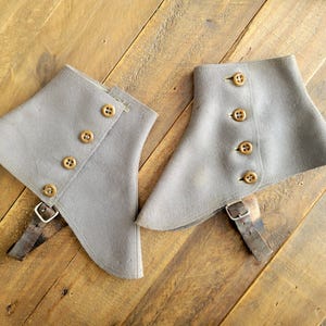 May include: A pair of gray spats with wooden buttons and leather straps. The spats have a curved design and are made of a soft, felt-like material. The buttons are evenly spaced down the front.