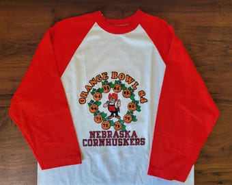 Könnte beinhalten: Ein Vintage-Raglan-Shirt mit weißem Körper und leuchtend roten Ärmeln. Das Shirt zeigt eine Grafik einer Cartoonfigur, umgeben von Orangen, sowie den Text "ORANGE BOWL '84" und "NEBRASKA CORNHUSKERS".