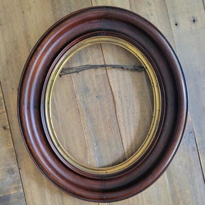 May include: A circular picture frame featuring a wide, dark brown wooden outer border with a smooth, rounded profile. The inner edge of the frame is lined with a thin, metallic gold-toned circular trim. A thin, dark wire is attached across the back.