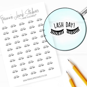 May include: A sheet of white sticker paper with black and white eyelash stickers that say "LASH DAY!"  The stickers are arranged in rows and columns. A magnifying glass is showing a close-up of one of the stickers.