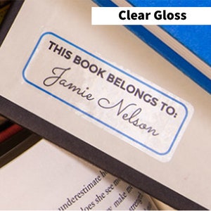 May include: Three labels with different finishes: clear frosty matte, clear gloss, and white matte. The labels are on various products, including spice jars, a book, and a candle. The text on the labels reads "Rosemary", "Garlic Powder", "This book belongs to Jamie Nelson", and "Lavender Vanilla".