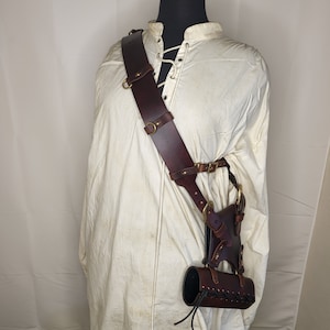 May include: A brown leather shoulder holster with a cylindrical pouch attached. The holster has brass accents and is worn over a white shirt.