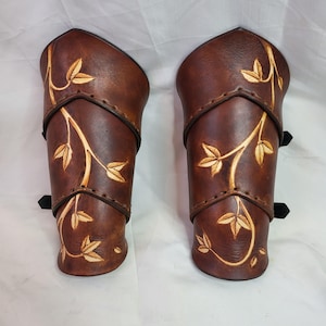 May include: A pair of brown leather arm guards with a vine and leaf design. The guards are made of thick leather and have a decorative stitching detail.