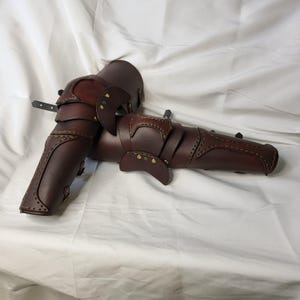 May include: A pair of brown leather knee pads with decorative stitching and metal accents. The pads are designed to protect the knees during activities such as LARPing or cosplay.