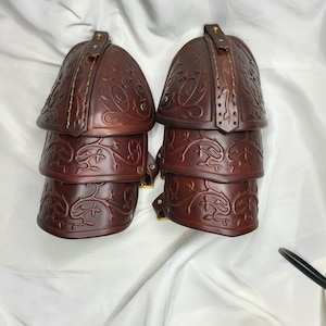 May include: A pair of brown leather arm guards with embossed floral patterns. Each guard features three articulated sections and a top piece with a strap and buckle. The leather is a deep brown, and the design is intricate.