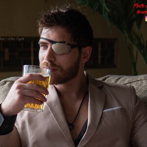 Peut inclure: Un homme portant un blazer beige et un cache-&oelig;il noir est assis sur un canap&eacute; et boit dans un verre de bi&egrave;re. Il porte un collier noir avec un pendentif circulaire. Le texte "Carbon Fiber - Killer Coyote" est visible dans le coin sup&eacute;rieur gauche de l'image.