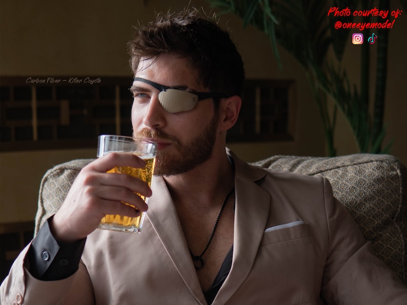 Peut inclure: Un homme portant un blazer beige et un cache-&oelig;il noir est assis sur un canap&eacute; et boit dans un verre de bi&egrave;re. Il porte un collier noir avec un pendentif circulaire. Le texte "Carbon Fiber - Killer Coyote" est visible dans le coin sup&eacute;rieur gauche de l'image.