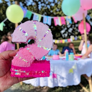 Peut inclure: Une piñata rose en forme de chiffre deux avec des accents irisés. La piñata est tenue devant une scène de fête avec des ballons et une table. La piñata est faite de franges de papier rose et magenta.