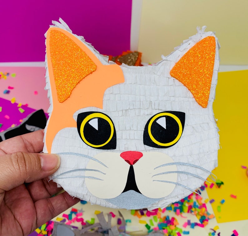 May include: A handmade cat-shaped pi&ntilde;ata with a white body, orange ears, and large black and yellow eyes. The cat's face has a pink nose and white cheeks. The pi&ntilde;ata is held by a hand, with colorful confetti in the background.