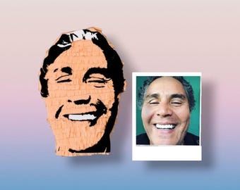 May include: A piñata in the shape of a smiling face with black outlines and a textured, light brown surface. Next to it is a framed photograph of the same smiling face.