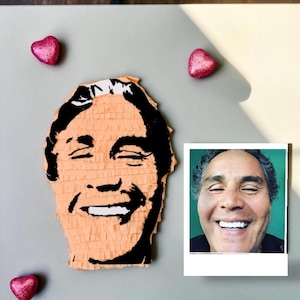 May include: A peach-colored pi&ntilde;ata shaped like a human head with black paper fringe detailing the facial features, accompanied by a square photograph of the same smiling person and three small, shiny, pink heart-shaped decorations.