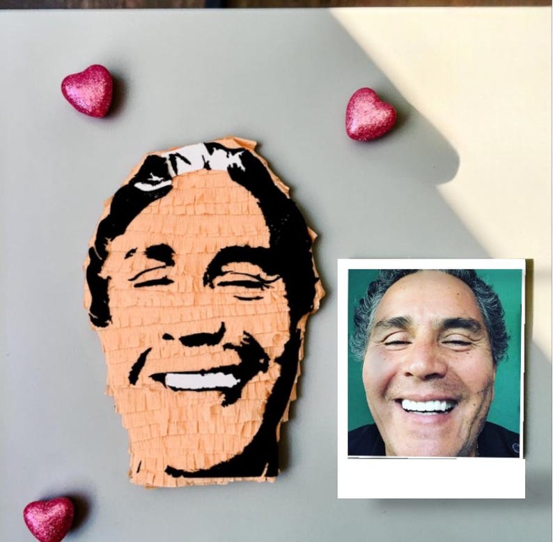May include: A peach-colored pi&ntilde;ata shaped like a human head with black paper fringe detailing the facial features, accompanied by a square photograph of the same smiling person and three small, shiny, pink heart-shaped decorations.