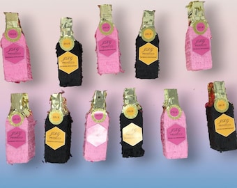 May include: An assortment of pink and black piñata bottles with gold foil tops. Each bottle has a label that reads "JRG Prosecco" and the year "2019". The piñatas are arranged on a gradient pink and blue background.