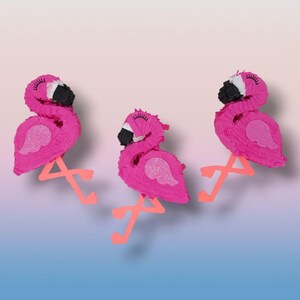 May include: Three pink flamingo piñatas with black beaks, white accents, and glittery wings. The flamingo legs are a bright orange color. The piñatas are arranged on a gradient background of pink and blue.