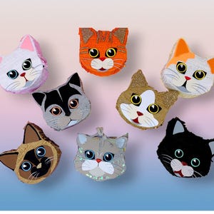 May include: A collection of colorful cat-shaped pi&ntilde;atas. Each pi&ntilde;ata features a unique design with different colors, including orange, gray, white, and black. The cat faces have large eyes, whiskers, and red mouths. Some have glitter accents.