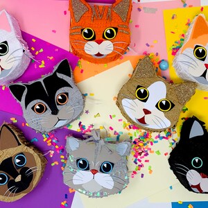 May include: A collection of cat-shaped pi&ntilde;atas in various colors, including white, orange, gray, and black. Each pi&ntilde;ata features detailed facial features and is decorated with glitter and confetti. A festive and colorful assortment.