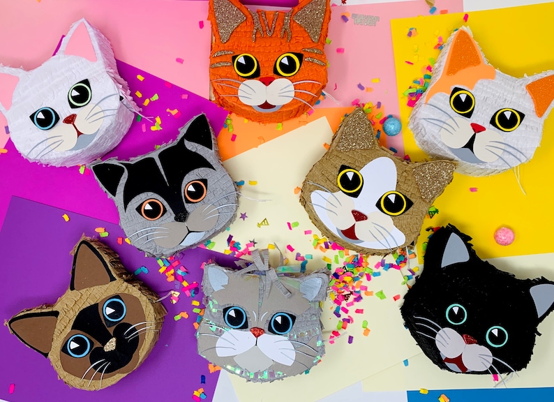 May include: A collection of cat-shaped pi&ntilde;atas in various colors, including white, orange, gray, and black. Each pi&ntilde;ata features detailed facial features and is decorated with glitter and confetti. A festive and colorful assortment.