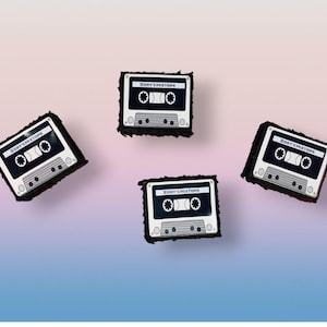May include: Four black and white cassette tape pins with the text "ZOBY'S MIXTAPE" on a gradient background. The pins have a retro design with a black border.