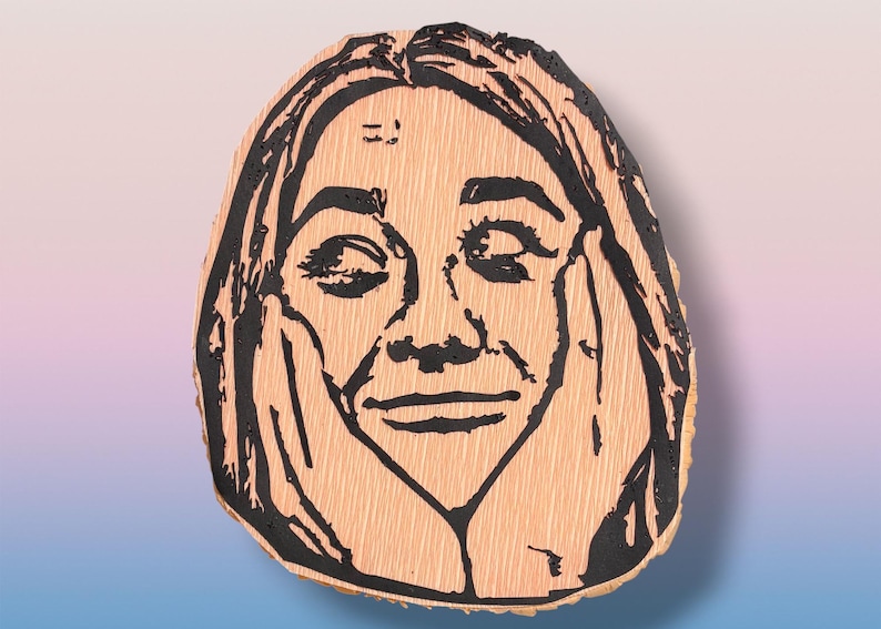 May include: A light-colored wooden slice featuring a black ink illustration of a person with long hair, eyes closed, and hands pressed against their cheeks. The wood grain is visible beneath the bold, dark lines of the portrait design.