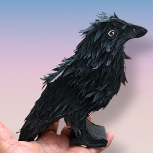 May include: A black raven figurine crafted with layered, textured paper feathers. The bird features a pointed beak, a circular eye with a white spiral design, and stands on a rectangular black base. The overall construction displays a detailed, feathered appearance.