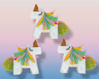 May include: Three white unicorn piñatas with colorful manes and tails. Each unicorn has a gold glitter horn and black eyelashes. The manes and tails feature rainbow colors, including yellow, green, and pink.