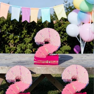 Puede incluir: Tres piñatas rosas con forma de número 2, con la parte superior rosa claro y la base rosa más oscura, decoradas con detalles brillantes. Una mesa de picnic de madera sostiene las piñatas, con globos de colores y una pancarta de fondo. Decoraciones de fiesta.