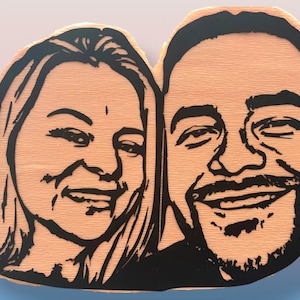 May include: A custom wooden plaque featuring a laser-cut or engraved black line art portrait of two people side-by-side. The light-colored wood grain is visible throughout the background, while the facial features are defined by bold black lines.