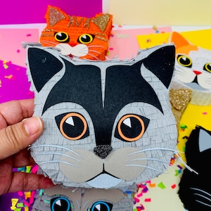 May include: A gray cat-shaped pi&ntilde;ata with black and orange accents is held in a hand. Other cat-shaped pi&ntilde;atas in orange, white, and black are visible in the background. The pi&ntilde;atas have large eyes and are decorated with glitter.