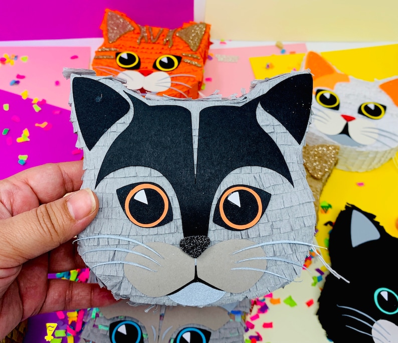 May include: A gray cat-shaped pi&ntilde;ata with black and orange accents is held in a hand. Other cat-shaped pi&ntilde;atas in orange, white, and black are visible in the background. The pi&ntilde;atas have large eyes and are decorated with glitter.