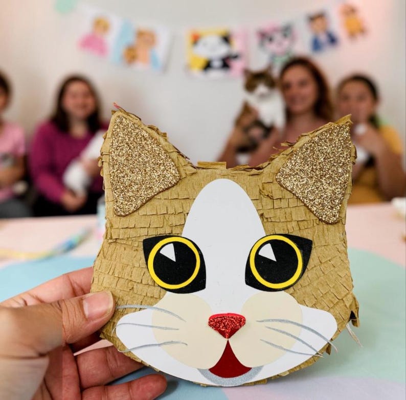 May include: A cat-shaped pi&ntilde;ata with a brown, white, and gold design. The ears are gold glitter, and the eyes are black with yellow outlines. The cat's nose is red glitter, and the mouth is open. The pi&ntilde;ata is held in front of a blurred background.