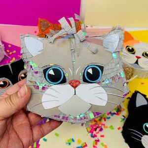 May include: A gray cat-shaped pi&ntilde;ata with large blue eyes, a red glitter nose, and white accents. The pi&ntilde;ata has a fringe of silver and iridescent paper. Other cat pi&ntilde;atas in various colors are visible in the background.