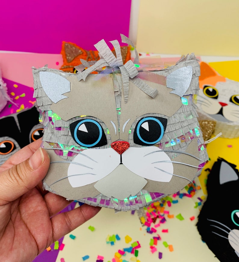 May include: A gray cat-shaped pi&ntilde;ata with large blue eyes, a red glitter nose, and white accents. The pi&ntilde;ata has a fringe of silver and iridescent paper. Other cat pi&ntilde;atas in various colors are visible in the background.