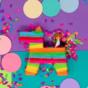 May include: A colorful, handmade piñata in the shape of a donkey, decorated with vibrant rainbow-colored fringe. Confetti and decorative circles in various colors surround the piñata, creating a festive party scene. The background is a mix of purple and teal.