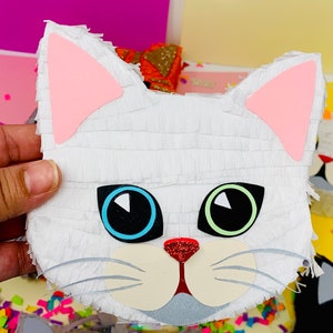 May include: A white cat-shaped pi&ntilde;ata with pink ears, blue and green eyes, and a red glitter nose. The pi&ntilde;ata is made of white crepe paper and has a whimsical design. The cat's face is the focal point of the pi&ntilde;ata.