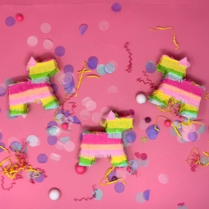 May include: Three colorful pinata shaped like animals, with pink, yellow, green, and blue fringe, on a pink background with confetti.