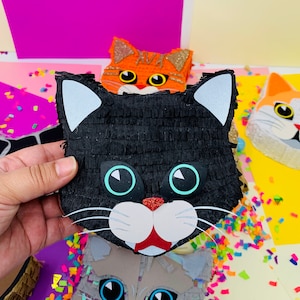 May include: A black cat pi&ntilde;ata with white ears, blue eyes, and a red nose is held in front of other cat-shaped pi&ntilde;atas. The pi&ntilde;ata is made of black crepe paper. Other cat pi&ntilde;atas are orange, gray, and white.