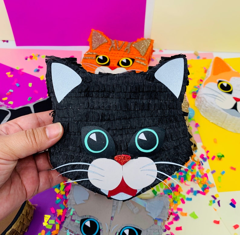 May include: A black cat pi&ntilde;ata with white ears, blue eyes, and a red nose is held in front of other cat-shaped pi&ntilde;atas. The pi&ntilde;ata is made of black crepe paper. Other cat pi&ntilde;atas are orange, gray, and white.